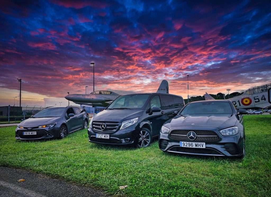 VTC Elvis Tres coches aparcados sobre el césped con un avión al fondo y un cielo de atardecer vibrante. servicios de VTC en Cantabria