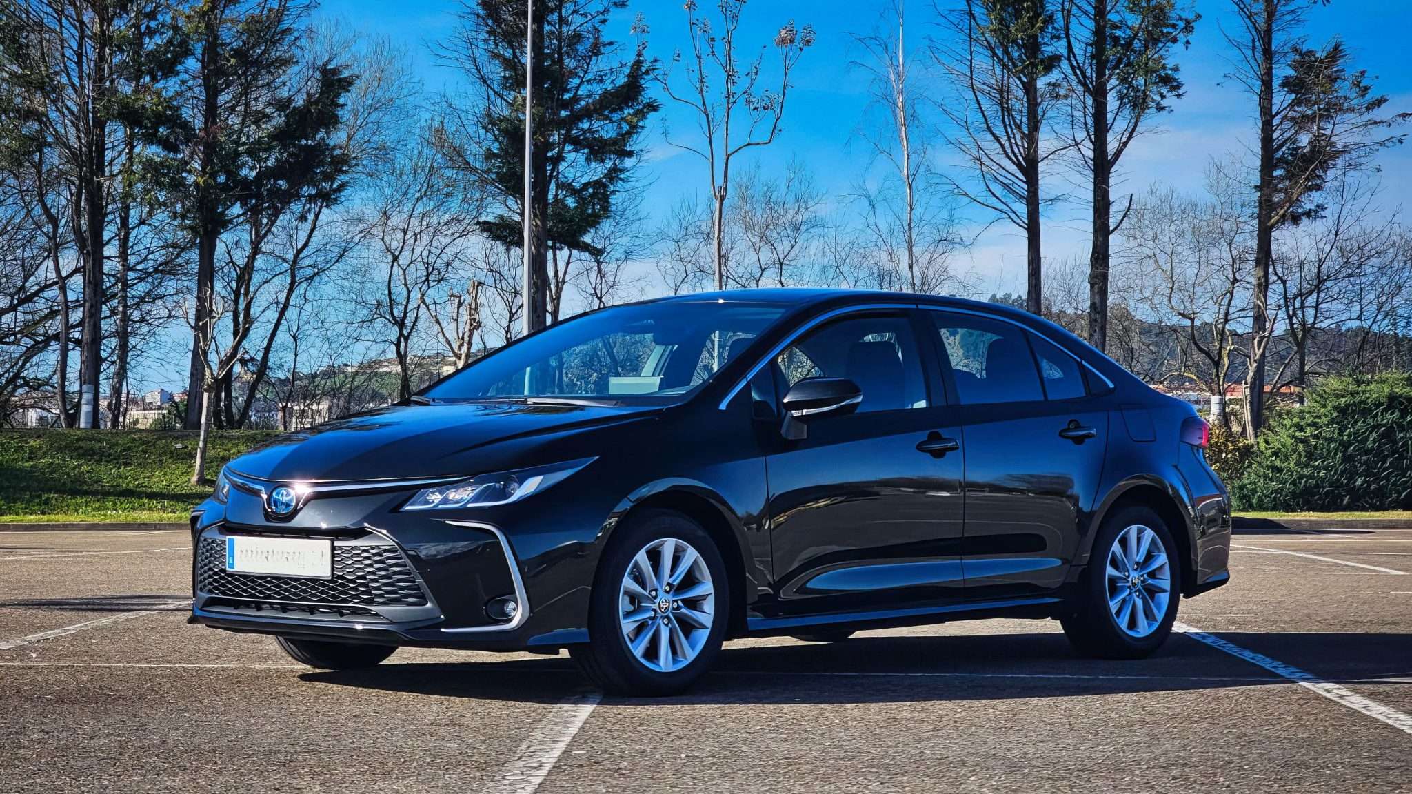 VTC Elvis Sedán negro aparcado en un solar vacío, listo para realizar tours turísticos en Cantabria, con árboles y un cielo azul al fondo. servicios de VTC en Cantabria