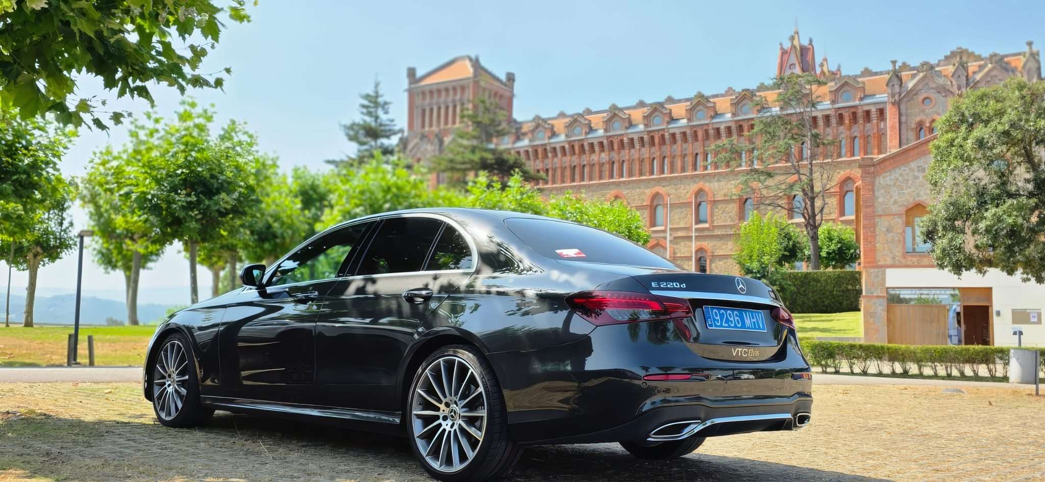VTC Elvis Un Mercedes-Benz sedán negro de una empresa de transporte privado en Cantabria aparcado junto a un edificio histórico de ladrillo en medio de una exuberante vegetación. servicios de VTC en Cantabria