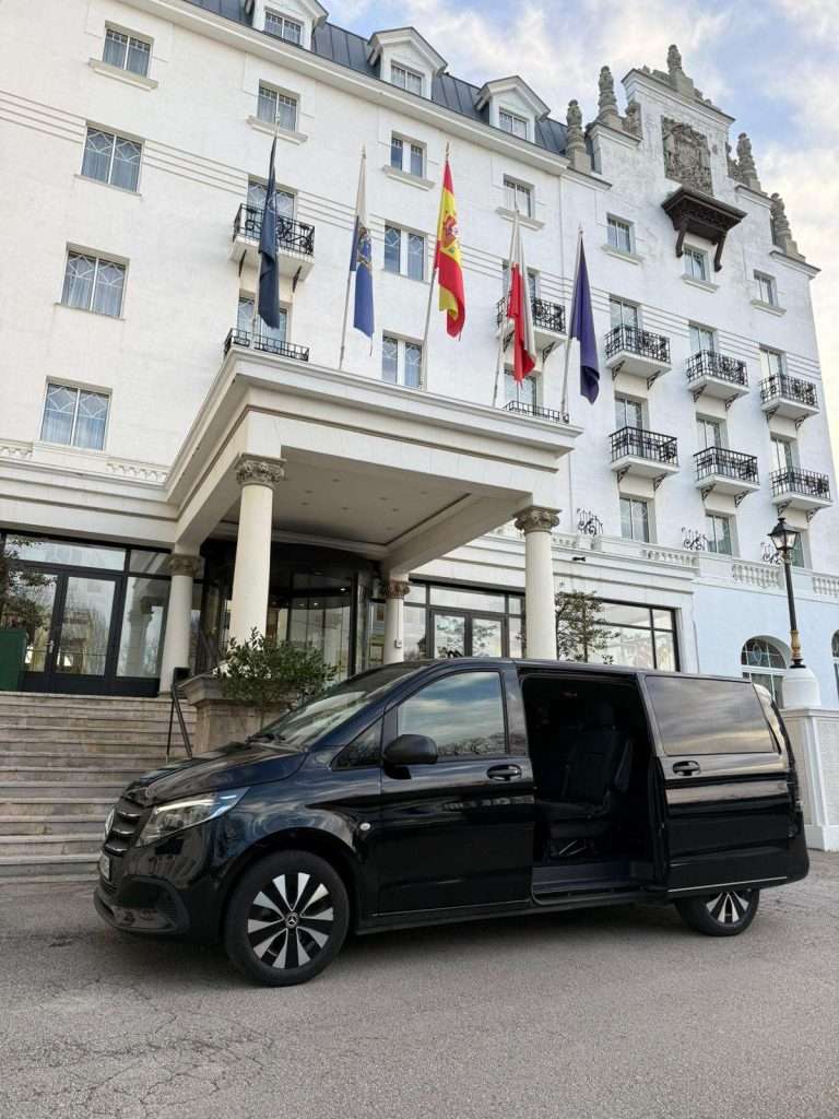 VTC Elvis Una furgoneta negra, utilizada para traslados al aeropuerto de Santander y Bilbao, está aparcada frente a un gran hotel blanco con banderas sobre la entrada. servicios de VTC en Cantabria