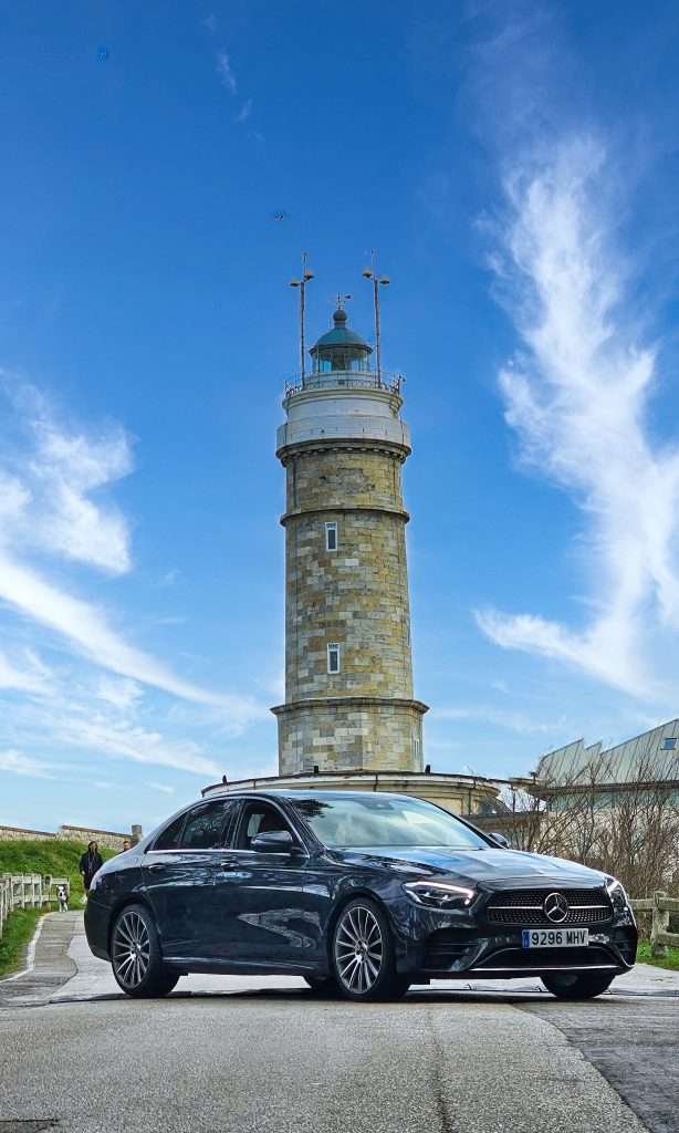 VTC Elvis Un Mercedes negro, listo para traslados a eventos privados, estacionado frente a un alto faro de piedra bajo un cielo azul con nubes tenues. servicios de VTC en Cantabria