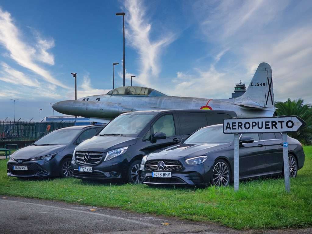 VTC Elvis Un día soleado arroja luz sobre una variedad de vehículos VTC, estacionados perfectamente junto a un cartel de aeropuerto con un avión antiguo como fondo, mostrando una flota impresionante. servicios de VTC en Cantabria