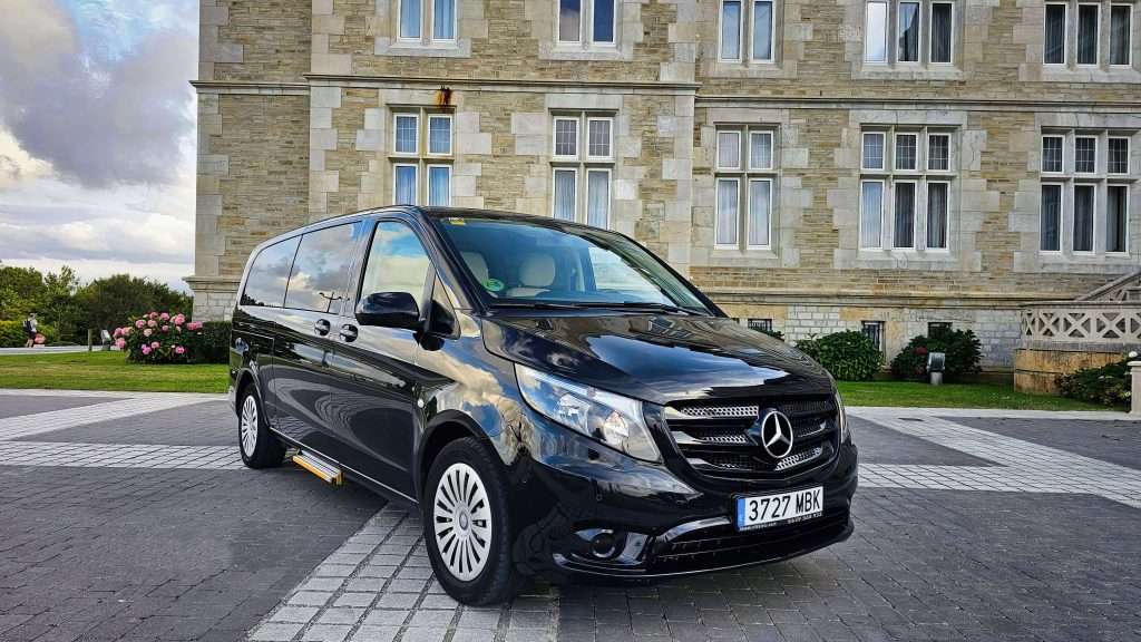 VTC Elvis Una furgoneta Mercedes-Benz de color negro, perfecta para el transporte de eventos privados en Cantabria, está aparcada delante de un edificio de piedra con grandes ventanales, con un cielo nublado como telón de fondo. servicios de VTC en Cantabria