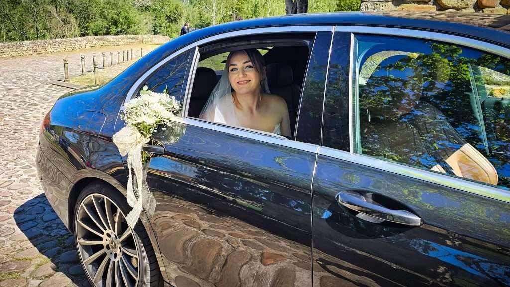VTC Elvis Una novia sonríe radiante dentro de un coche bellamente decorado, adornado con un ramo de flores en la puerta, estacionado en una calle adoquinada rodeada de exuberante vegetación: una escena ideal para coches para bodas en Cantabria. servicios de VTC en Cantabria