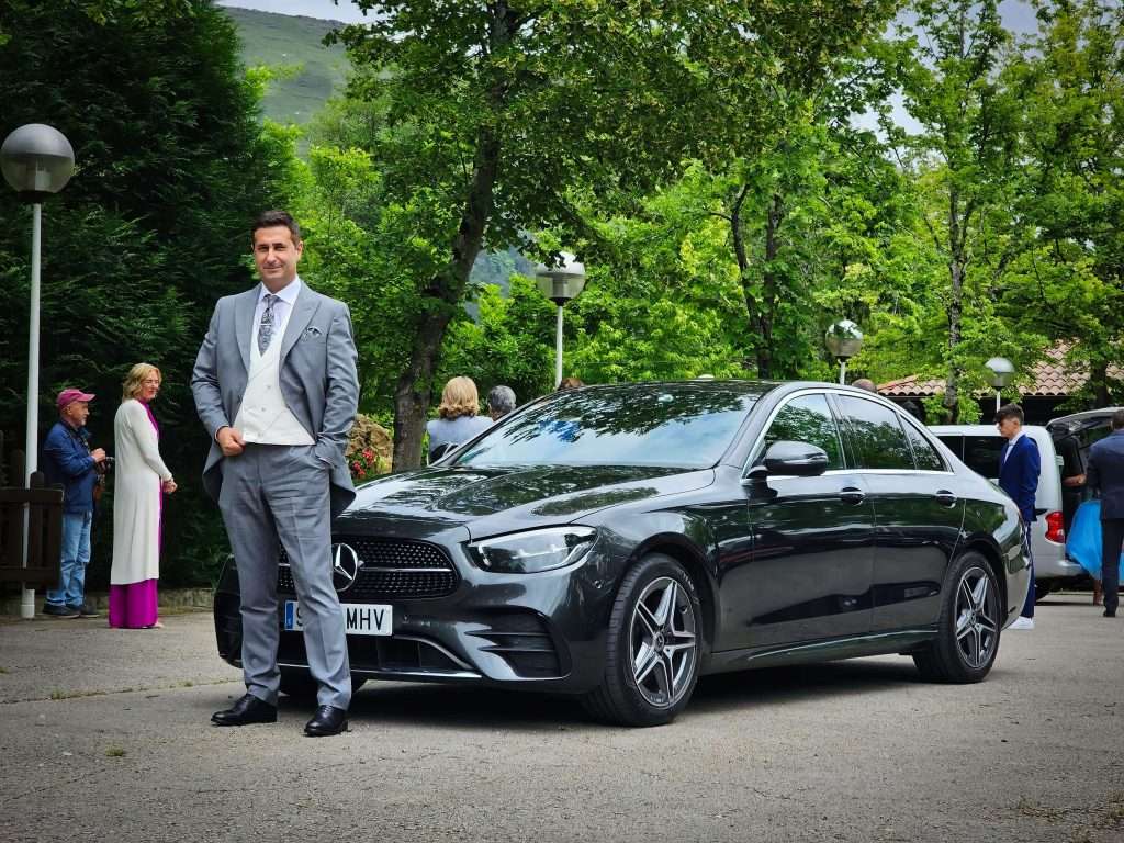 VTC Elvis Un hombre con un traje gris se encuentra junto a un elegante automóvil negro, ideal para ocasiones especiales como bodas en Cantabria, rodeado de árboles y gente mezclándose en el fondo. servicios de VTC en Cantabria