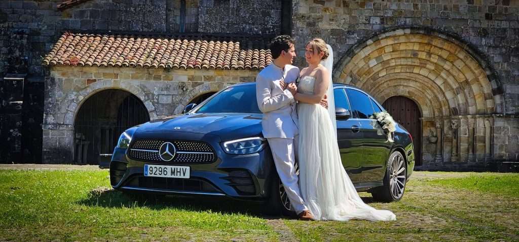 VTC Elvis Los novios, vestidos de gala, se encuentran junto a un Mercedes-Benz negro, símbolo de los coches para bodas en Cantabria, con un histórico edificio de piedra al fondo. servicios de VTC en Cantabria