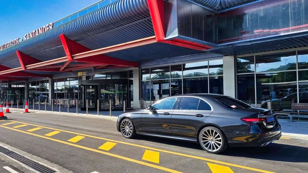 VTC Elvis Un elegante sedán negro, adornado con elegantes detalles en rojo, espera su próximo viaje bajo el cielo azul claro, listo para trasladarse al aeropuerto de Cantabria. La moderna terminal se alza como telón de fondo de esta combinación perfecta de estilo y funcionalidad. servicios de VTC en Cantabria