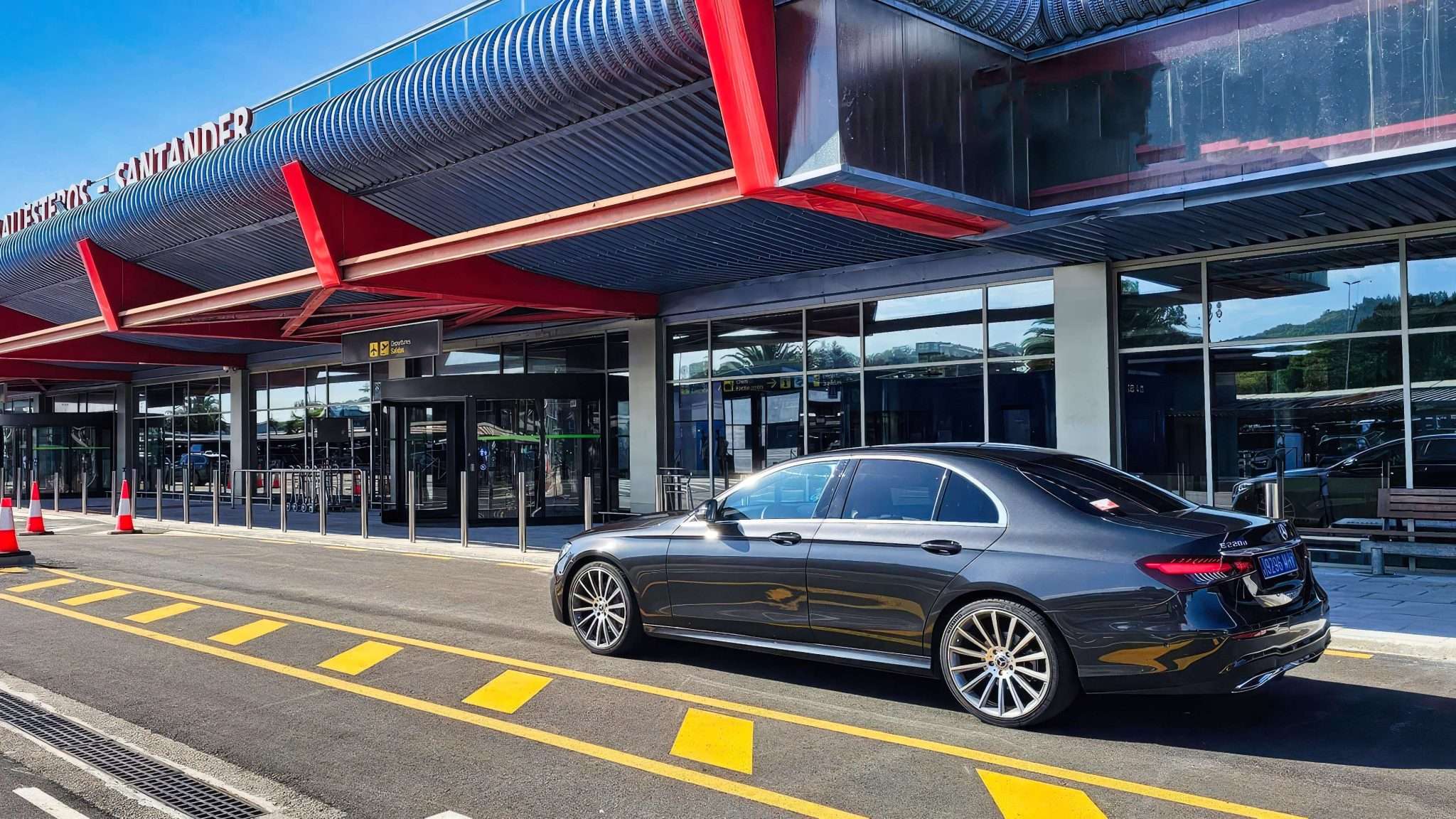 VTC Elvis Un elegante sedán negro, adornado con elegantes detalles en rojo, espera su próximo viaje bajo el cielo azul claro, listo para trasladarse al aeropuerto de Cantabria. La moderna terminal se alza como telón de fondo de esta combinación perfecta de estilo y funcionalidad. servicios de VTC en Cantabria