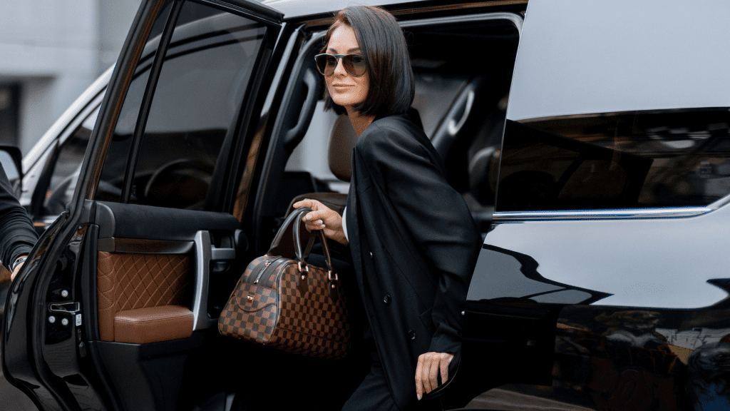VTC Elvis Una persona con gafas de sol sale elegantemente de un coche, luciendo un bolso a cuadros, personificando la elegancia de los artistas en traslados de Cantabria. servicios de VTC en Cantabria