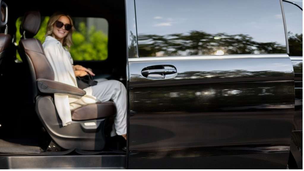 VTC Elvis Una mujer con gafas de sol está sentada en un coche con la puerta abierta, sonriendo, su ropa de color claro refleja la luz del día. Los árboles se reflejan de forma hermosa en el exterior del coche, lo que sugiere viajes tranquilos por Cantabria durante los traslados de los artistas. servicios de VTC en Cantabria