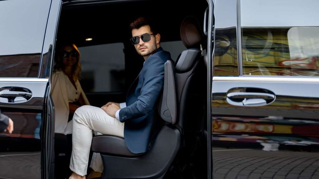 VTC Elvis En el asiento trasero de un coche con la puerta abierta, un hombre con gafas de sol se sienta junto a una mujer. Probablemente estén disfrutando de uno de esos exclusivos traslados de artistas en Cantabria, que combinan a la perfección comodidad y estilo en su viaje. servicios de VTC en Cantabria