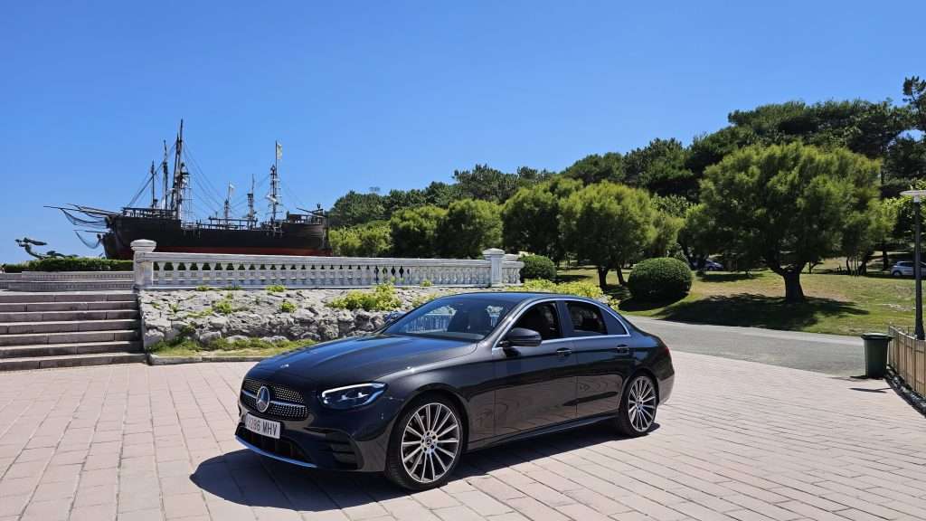 VTC Elvis Un elegante Mercedes-Benz negro, a menudo utilizado para traslados VIP para famosos, está estacionado en un área pavimentada con un barco histórico y una exuberante vegetación en el fondo bajo un cielo azul claro. servicios de VTC en Cantabria