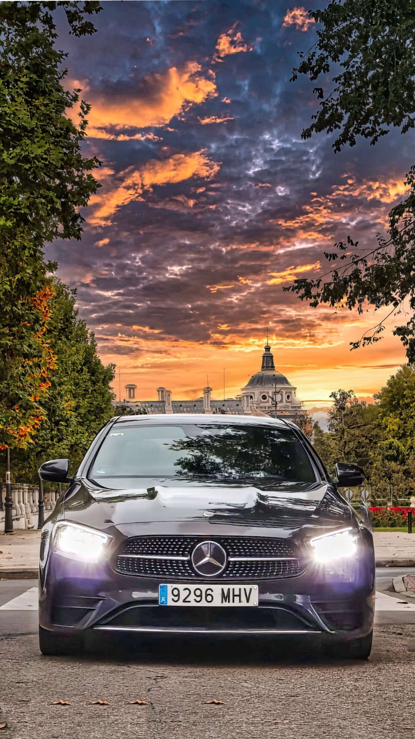 VTC Elvis Un elegante automóvil Mercedes-Benz gris, con sus faros iluminando la calle, está estacionado frente a un fondo de una vibrante puesta de sol y un edificio antiguo. servicios de VTC en Cantabria