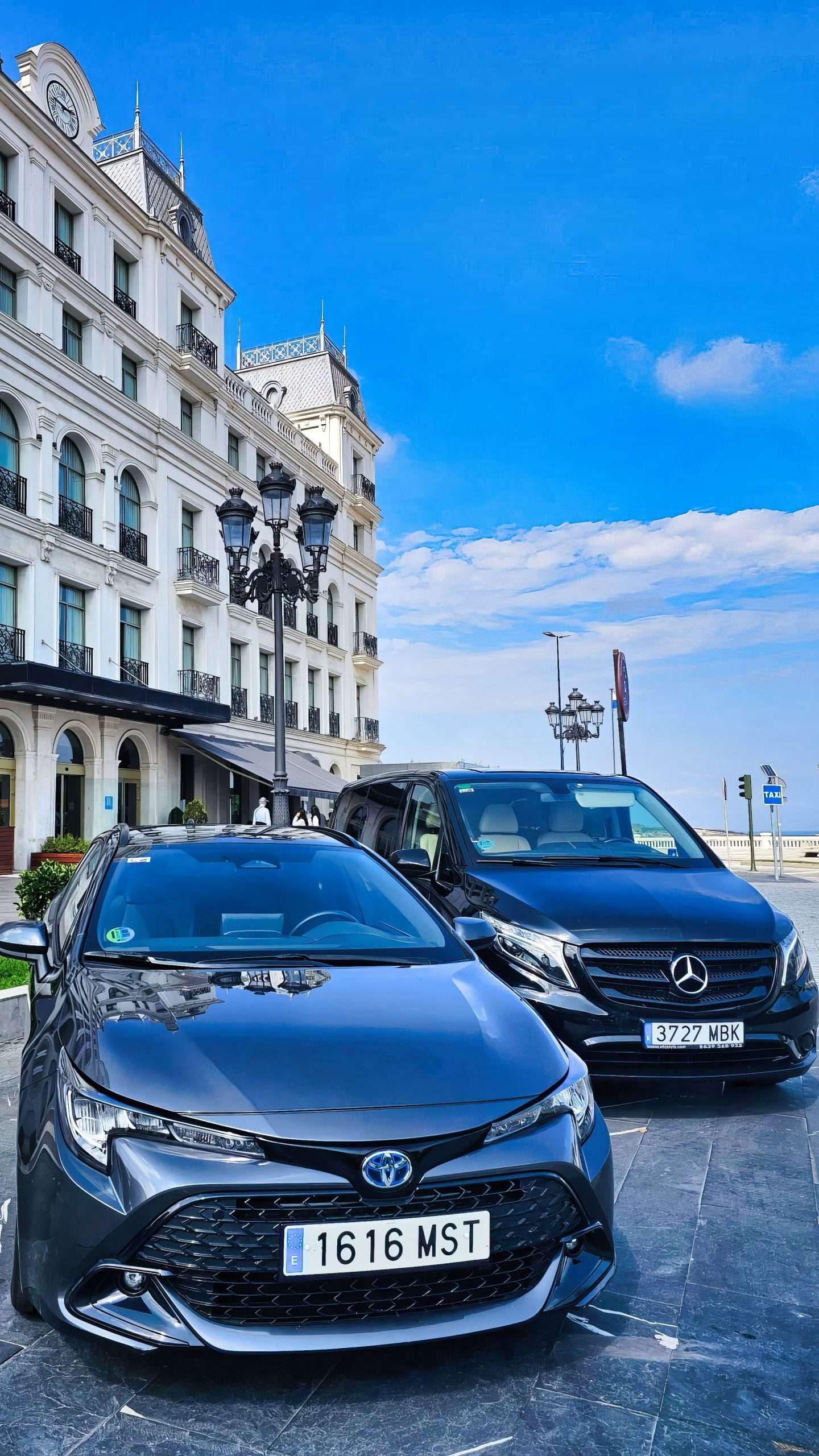 VTC Elvis Un Toyota negro y un Mercedes negro estacionados frente a un elegante edificio con una arquitectura ornamentada bajo un cielo azul claro. servicios de VTC en Cantabria