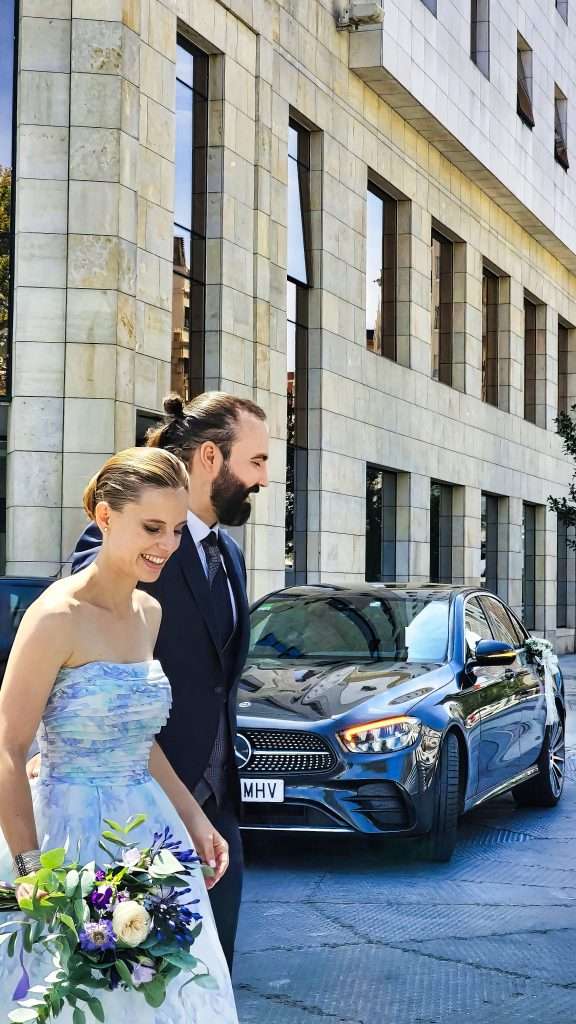 VTC Elvis Una pareja vestida formal sonríe y camina junta, con un ramo de flores en la mano, cerca de un edificio moderno y un automóvil Mercedes estacionado, mientras disfrutan de un traslado perfecto para bodas en su día especial. servicios de VTC en Cantabria