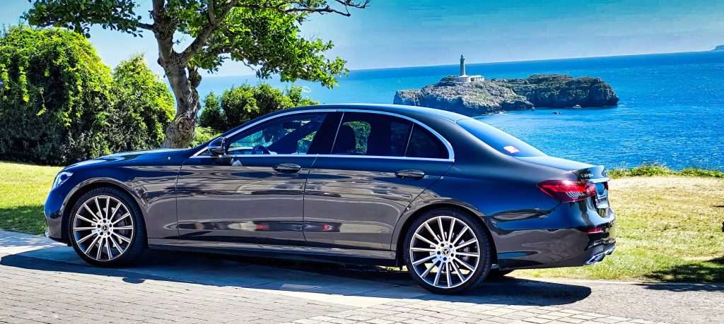 VTC Elvis Un elegante sedán negro, parte de los principales servicios de traslados al hotel, está estacionado junto al mar con una isla y un faro al fondo bajo un cielo azul claro. servicios de VTC en Cantabria