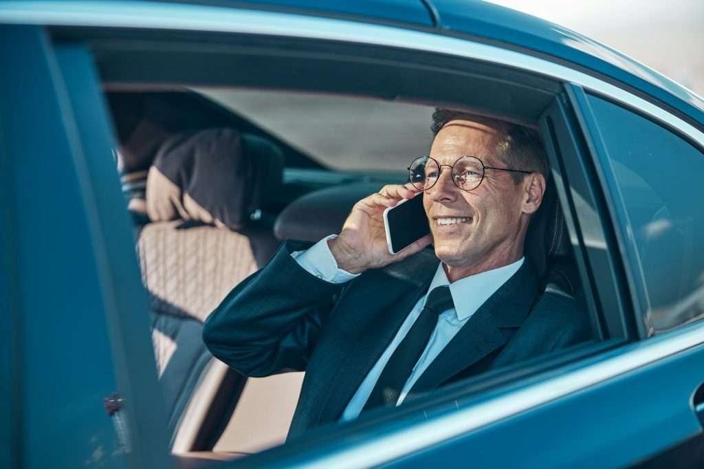 VTC Elvis Un hombre con traje y gafas está sentado en un coche, sonriendo y hablando por un teléfono inteligente. servicios de VTC en Cantabria