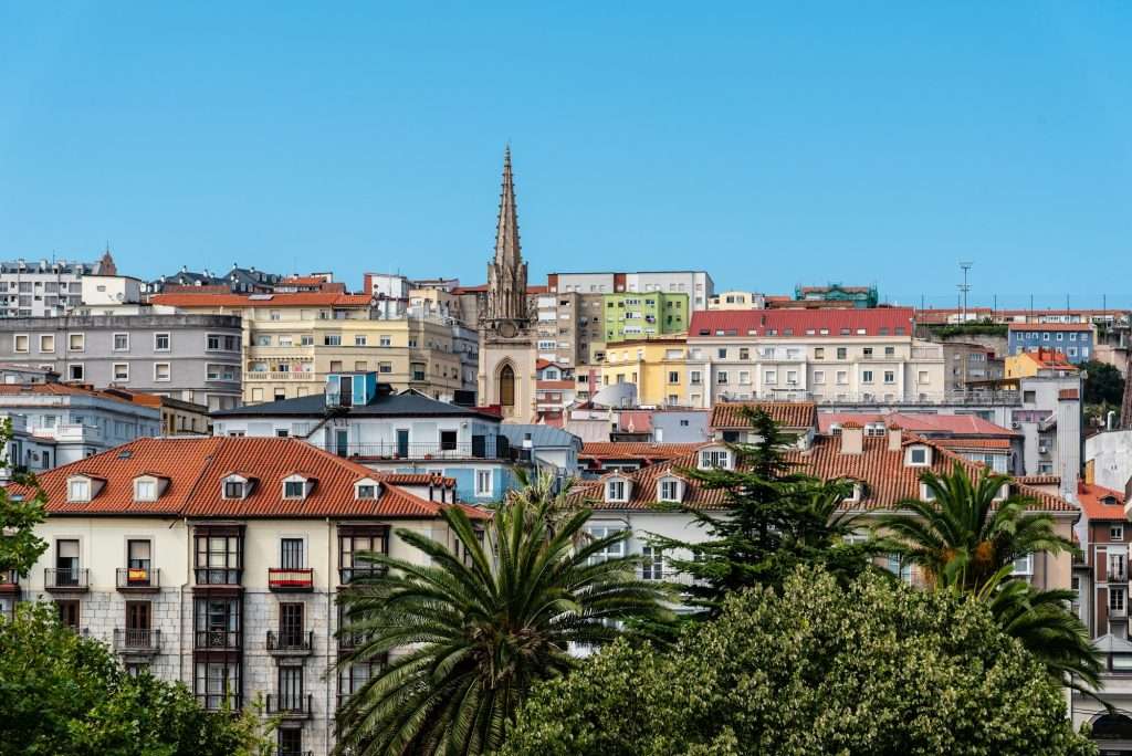 VTC Elvis Vista panorámica de una ciudad con edificios coloridos, una torre de iglesia y exuberante vegetación bajo un cielo azul claro. servicios de VTC en Cantabria