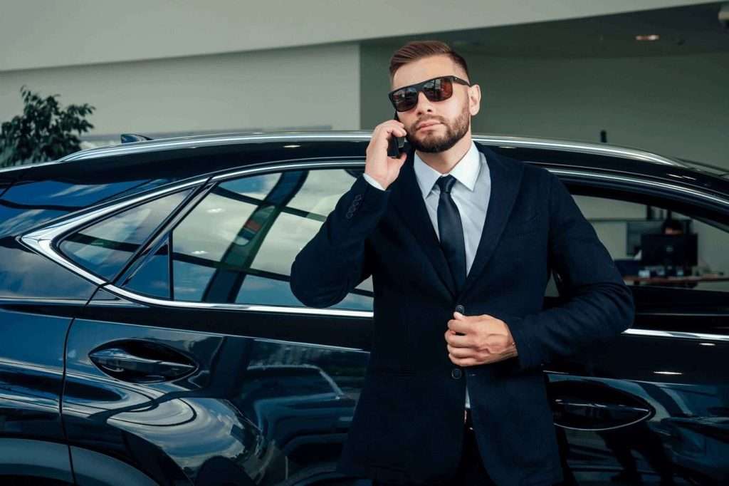 VTC Elvis Hombre con traje y gafas de sol, hablando por un teléfono celular, de pie junto a un automóvil negro dentro de una sala de exposición moderna. servicios de VTC en Cantabria